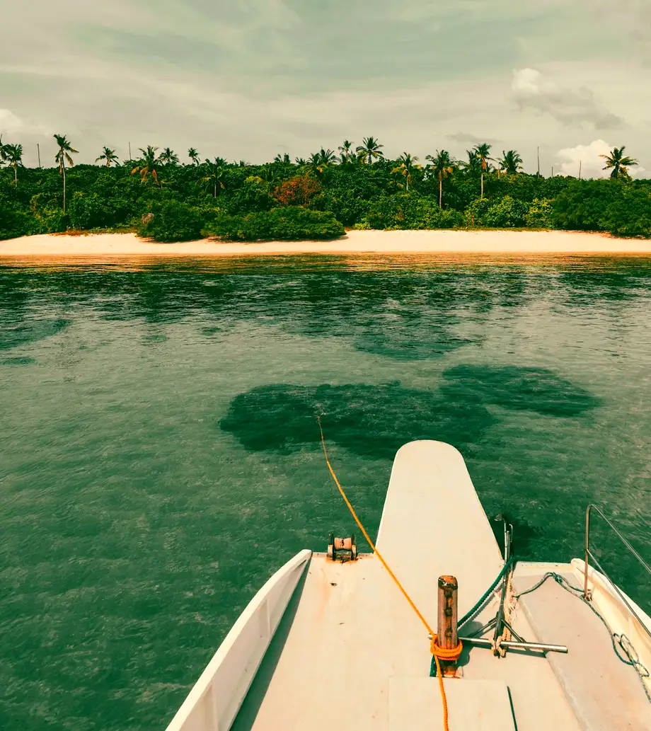 The bow of a boat pointing across clear shallow water toward a palm-lined white beach
