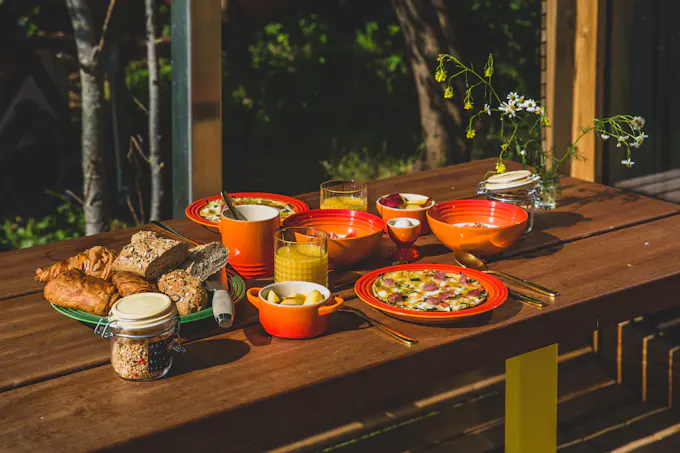 A wooden table outdoors set with bread, fruit, juice and coffee in orange dishes