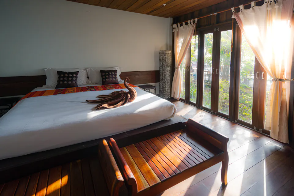 A wooden guest room in warm morning light, glass-paned doors opening onto green trees