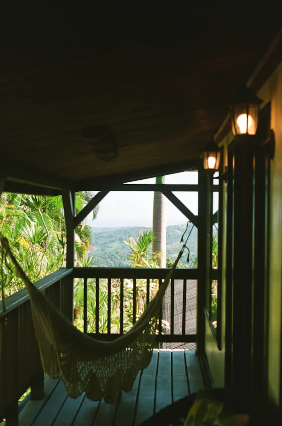 A woven hammock hanging on a wooden veranda, palm trees and hills beyond the rail
