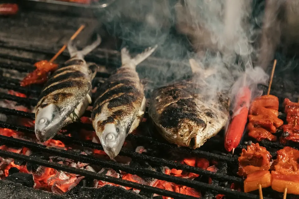 Three whole fish and skewers cooking on a grill over glowing coals, smoke rising