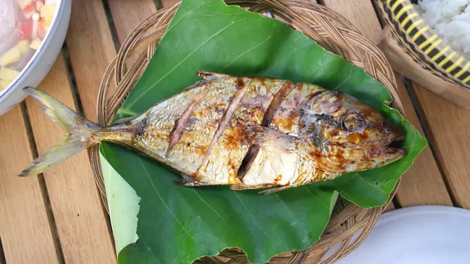 A whole grilled fish scored and glazed, resting on a banana leaf in a woven tray